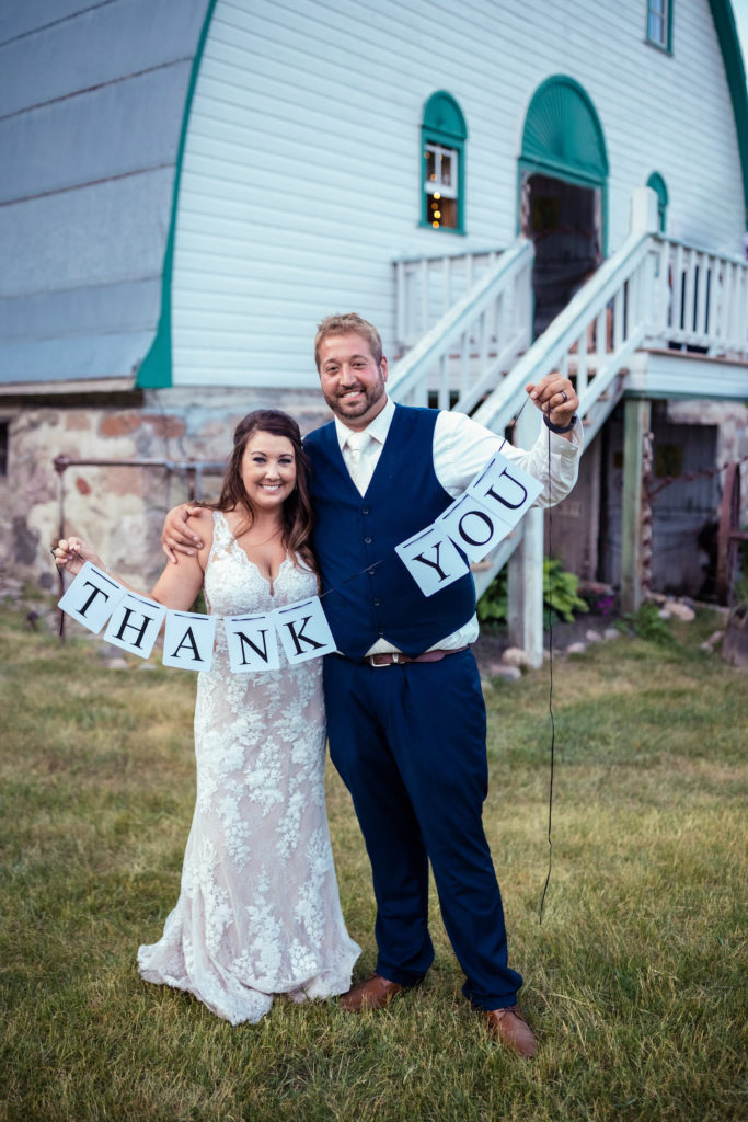 Bride and groom smile and pose in front of the ranch wedding location while holding up a banner that reads Thank You