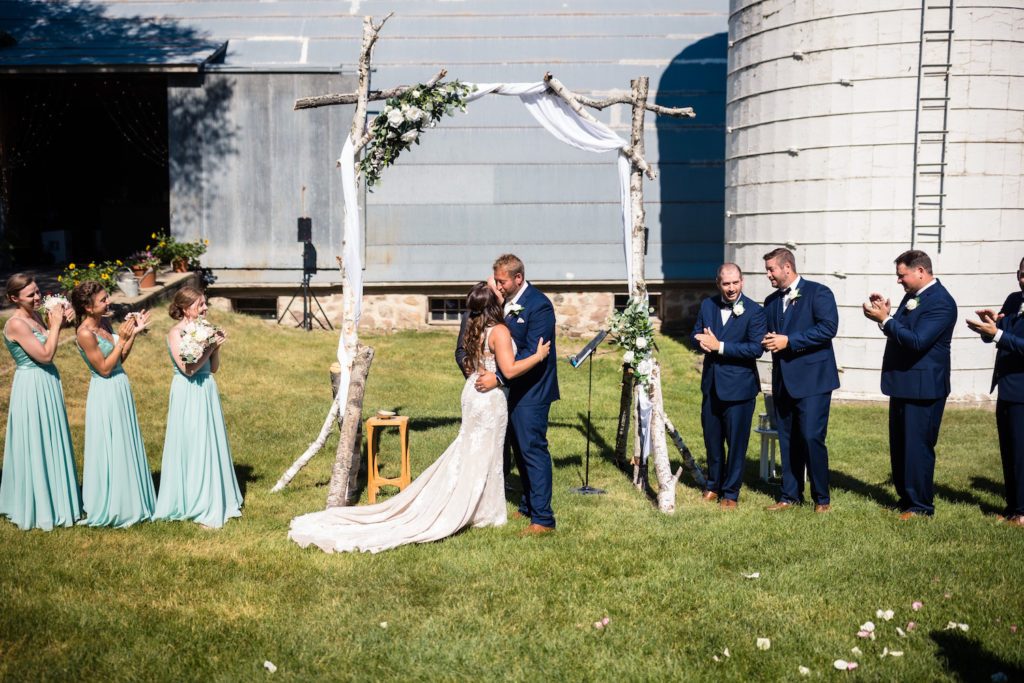 Bride and groom share a kiss under a makeshift wedding arch in front of a ranch as their guests look on