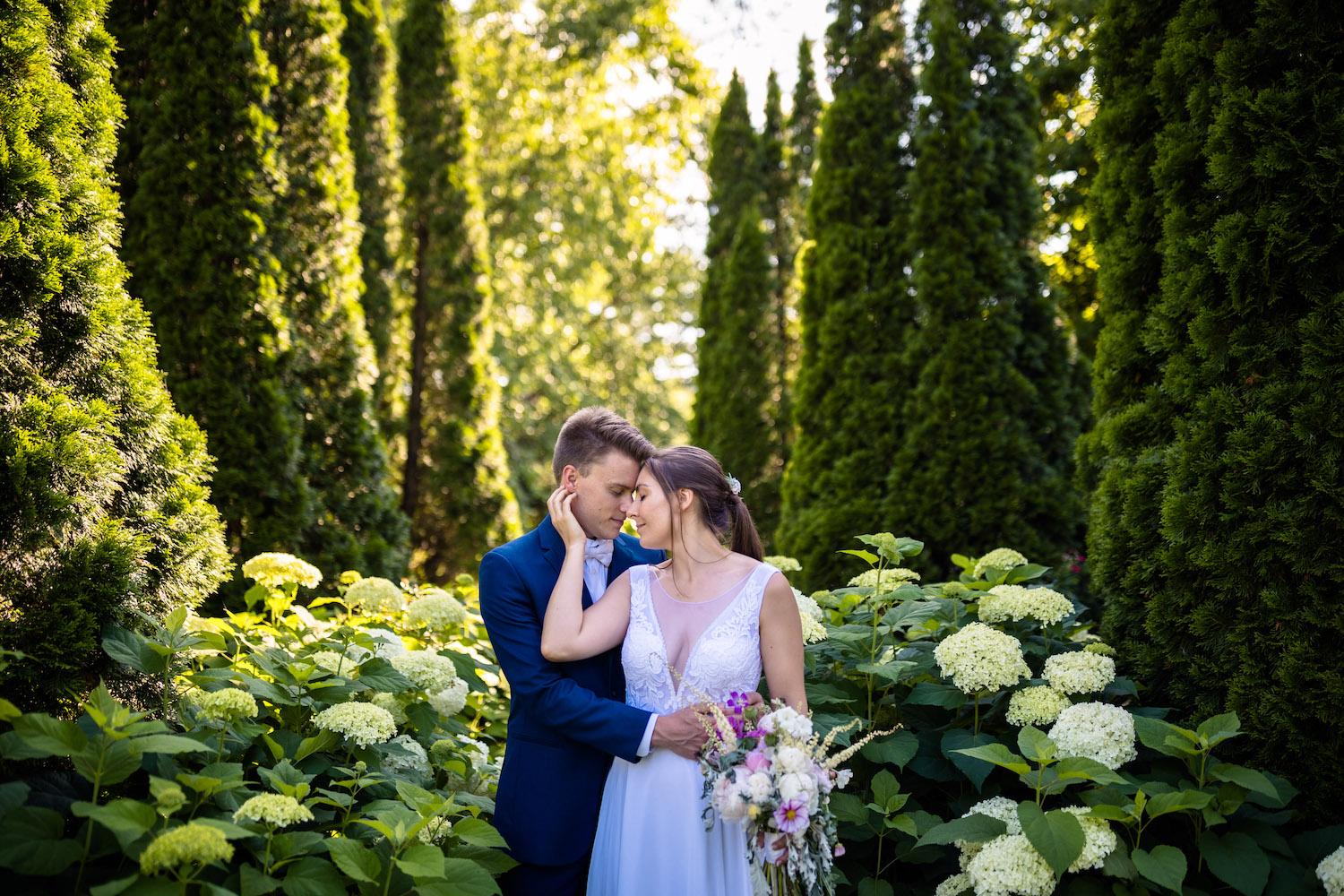 Bride and groom embrace and pose in front of elegant trees in a garden during their fairytale Lake Morey Resort wedding in Vermont by Makayla McGarvey