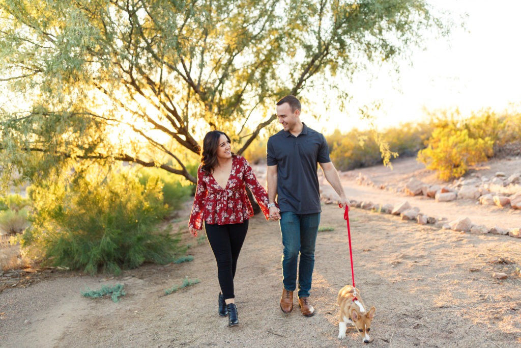 Couple holds hands as they smile at each other, with the guy holding the leash to their adorable dog, during their engagement session