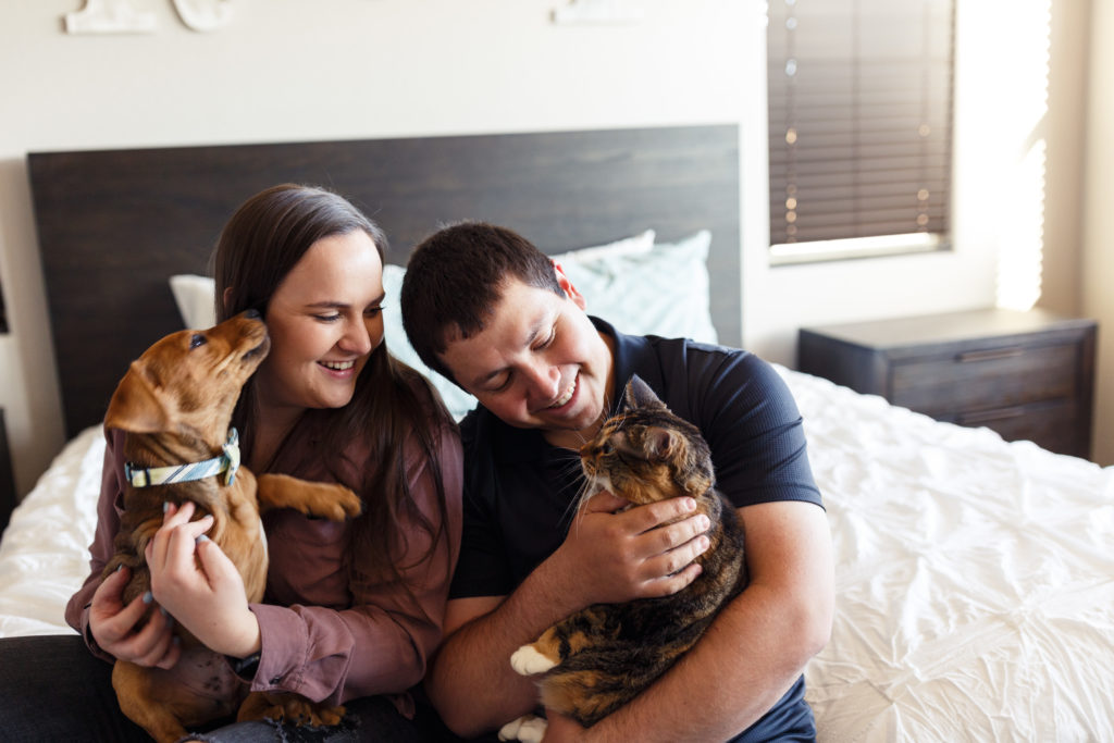 Couple holds on to their pets, with the girl holding their dog and the guy holding their cat, during their engagement photoshoot with pets in the bedroom