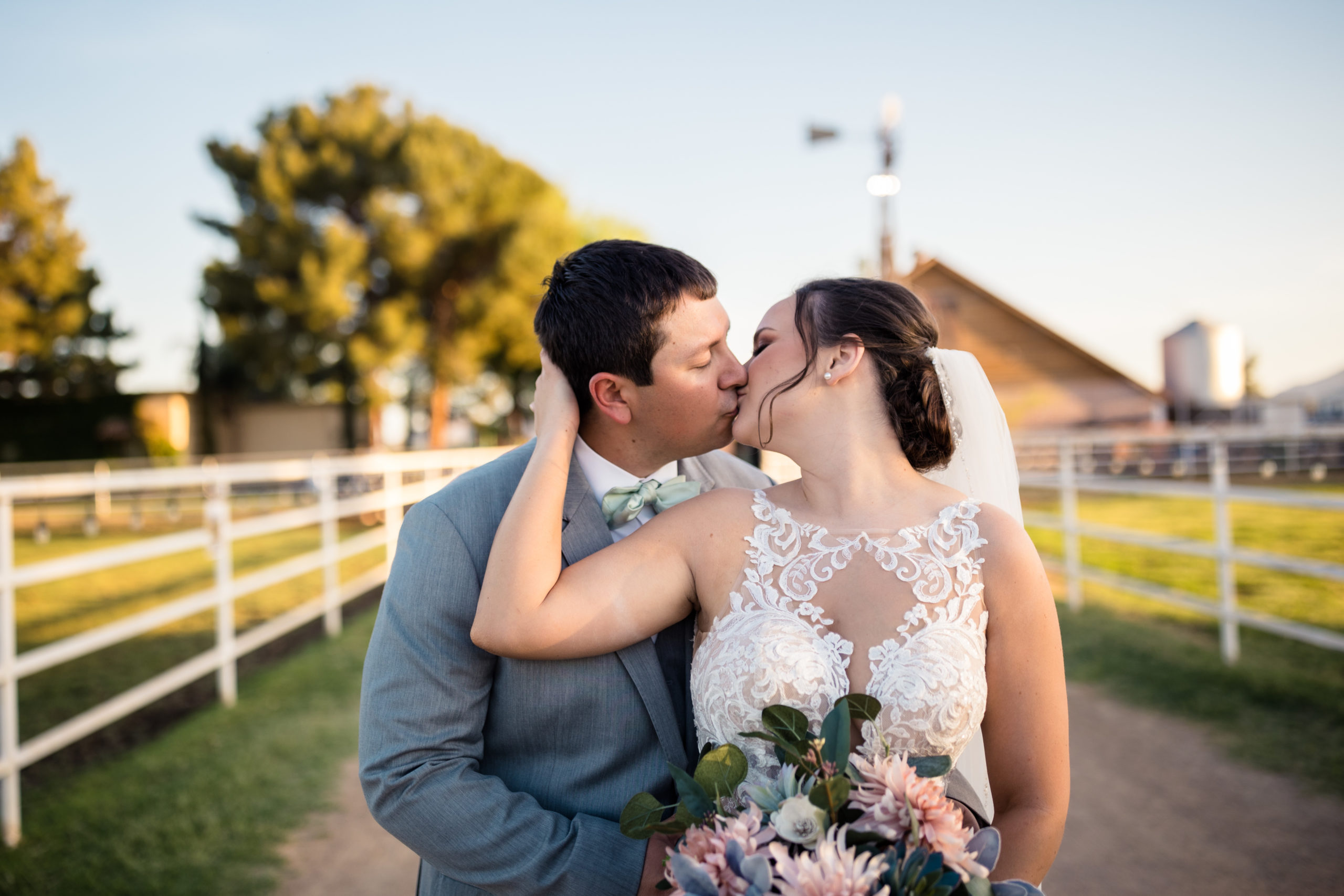 Bride places her hands on the back of the groom’s face as they share a kiss in front of the ranch and horse pens at The Knotty Barn, shot by Makayla MacGarvey