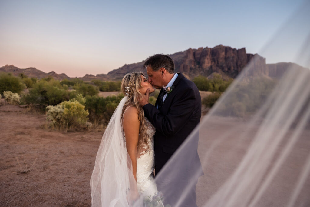 A couple is standing in front of Flatiron Rock in the Superstition Mountains on their elopement day. The groom is tilting the ride's chin up to kiss her.