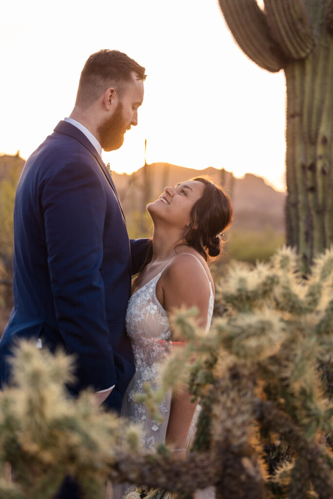 A couple is standing side by side in their elopement attire, in front of a tall Saguaro Cactus in the Superstition Mountains.