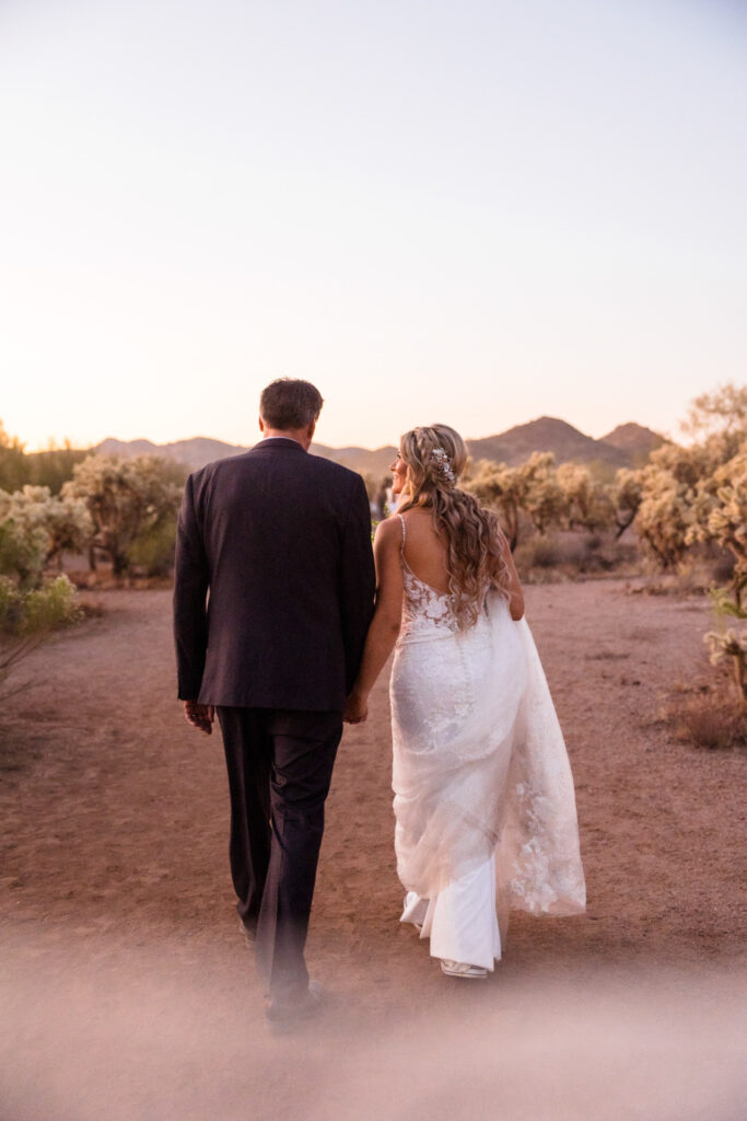 A couple is walking away, holding hands, dressed in wedding attire. They're celebrating their elopement in the Superstition Mountains!
