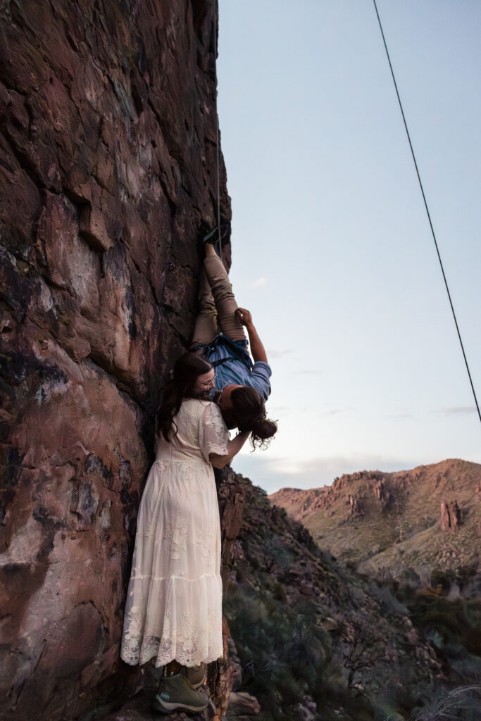 A bride standing at the bottom of the Totem Pole rock climbing location, while the groom hangs upside down to kiss her.