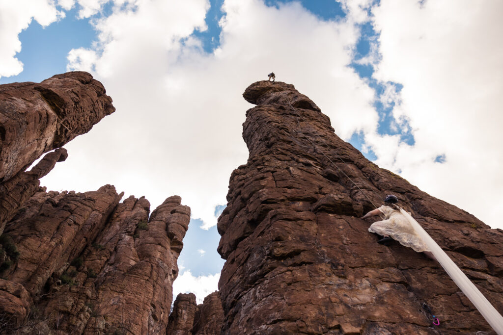 A couple rock climbing at Totem Pole in Arizona, wearing their wedding attire.