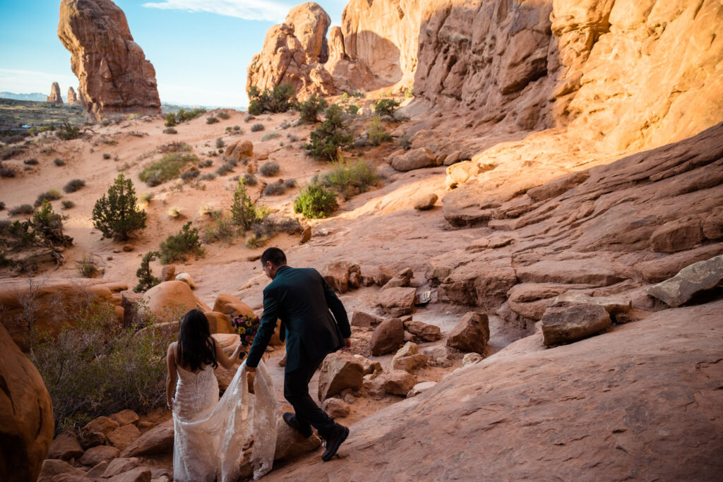 A couple walking on the sandstone at Arches National Park in their elopement attire. The groom is holding the bride's dress as they walk away.