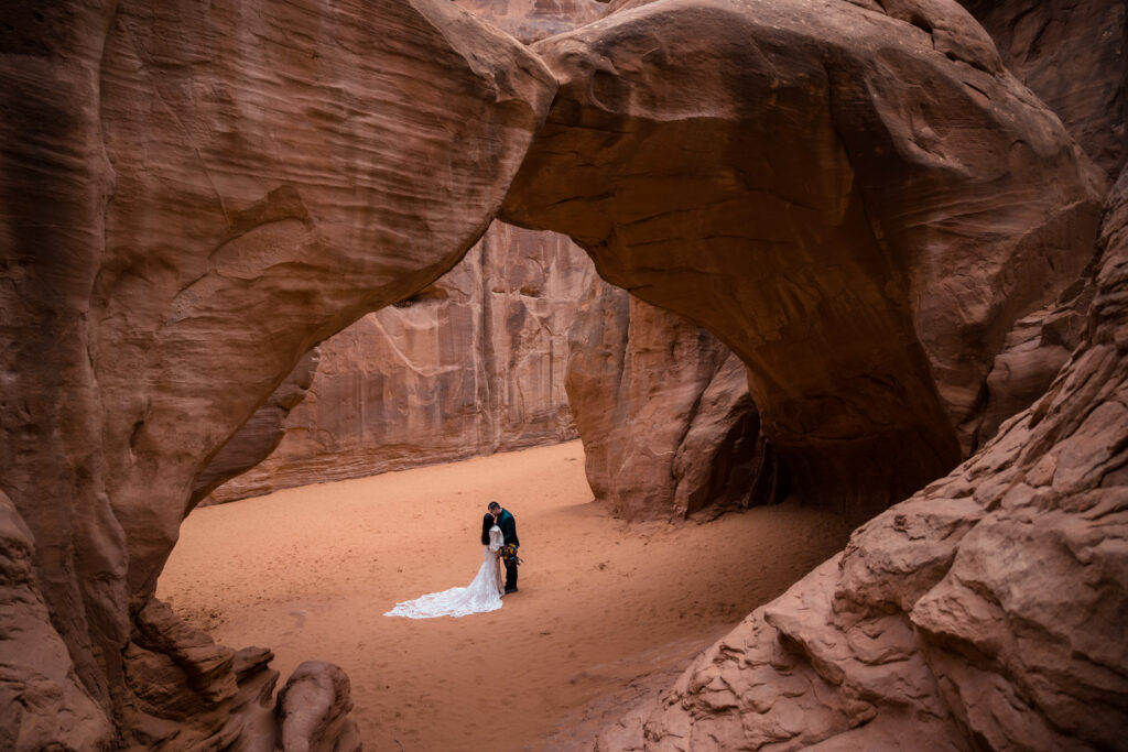 A couple standing under Sandstone Arch in Arches National Park after their elopement ceremony. They are facing each other and kissing, with the arch above them.