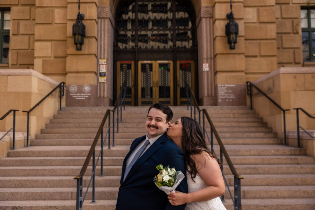 A couple standing outside the old Phoenix courthouse, at the bottom of the stairs on their wedding day. The bride is kissing the groom on the cheek.