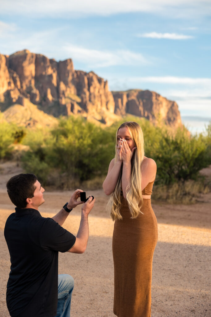 One of the best places to propose in Phoenix is the Superstition Mountains. A man is on one knee holding a ring, while his partner covers her face in shock.